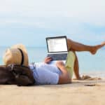 Man reading email on a laptop while relaxing on the beach, reflecting the self-employed lifestyle tied to Schedule SE.