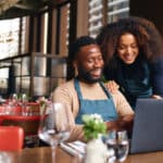 Small business owners using a laptop in a restaurant, reviewing income and adjustments that may be reported on IRS Schedule 1.