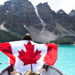 Young woman in a canoe holding a Canadian flag on Moraine Lake, symbolizing the adventure of Americans moving to Canada.