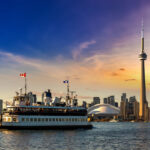 Panoramic view of the Toronto skyline and CN Tower at sunset, with a ferry crossing the water—showing city life and the cost of living in Canada.